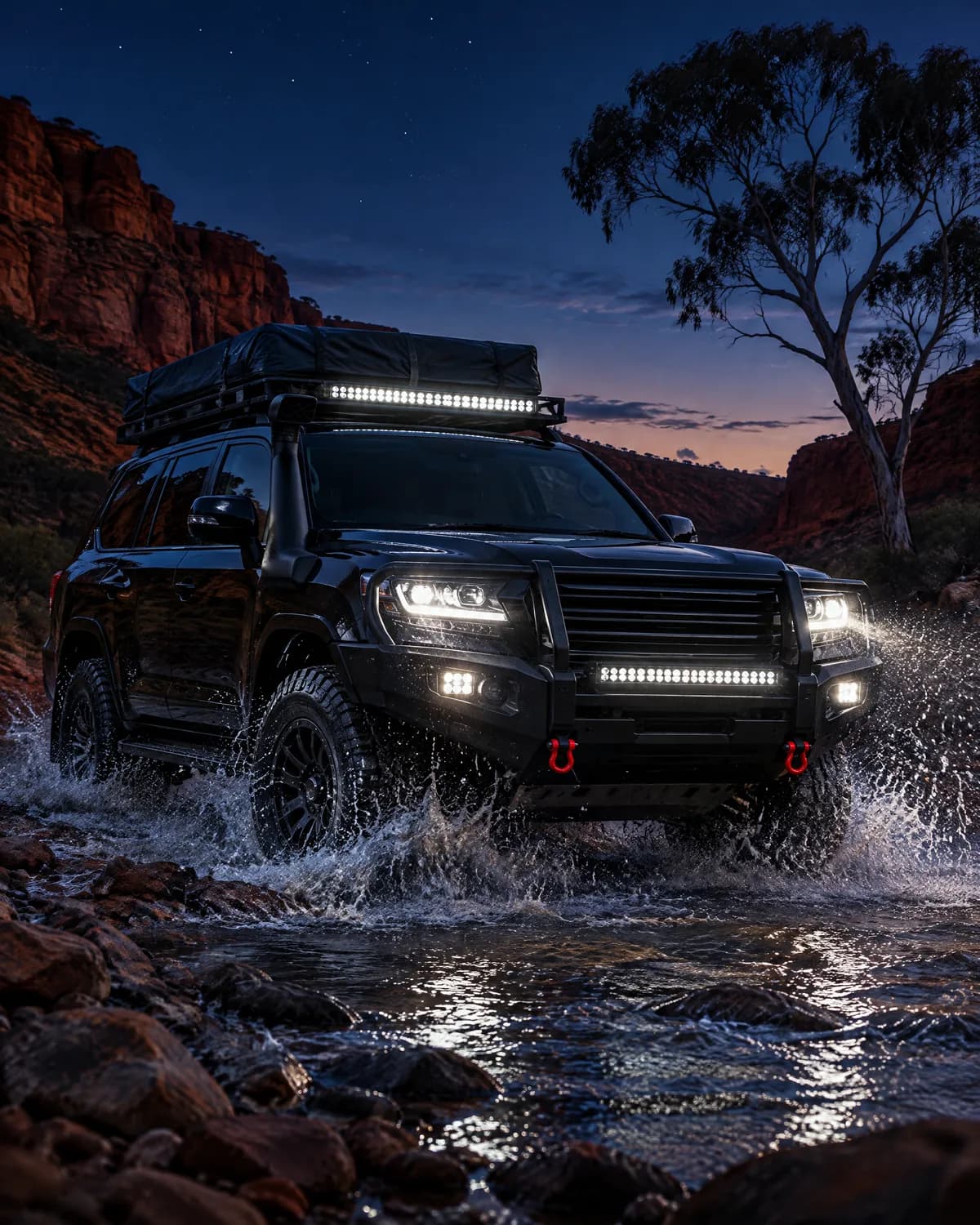 Black 4x4 touring wagon with a rooftop tent, light bars and red recovery shackles splashing through a rocky creek in a red sandstone gorge at dusk, a ghost gum beside the track.